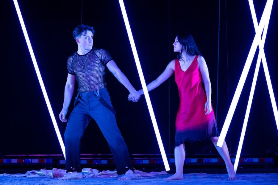 Two individuals with serious expressions walk between hanging rods of LED lights while holding hands. One of the individuals wears a red dress and the other individual wears a black mesh shirt and black pants.