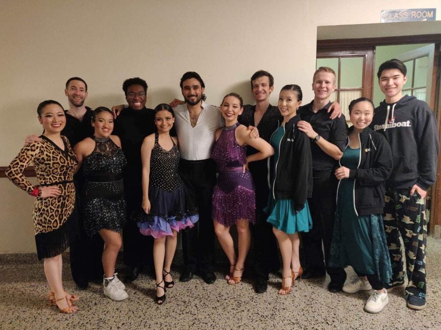 12 dancers in costumes and black jackets pose together.