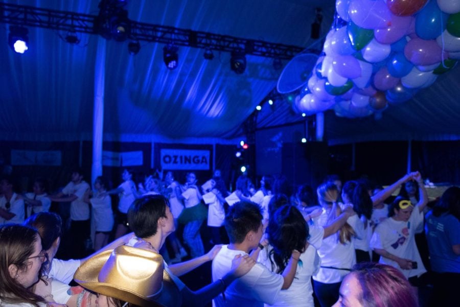 Students in white T-shirts dance in a conga line in a dark tent with blue lighting under a suspended mesh bag of multi-colored balloons.