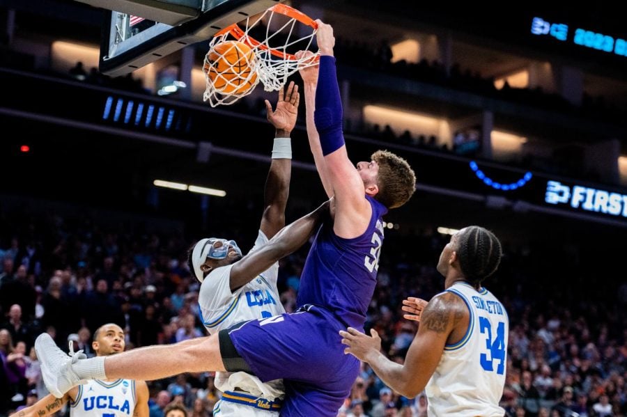 A player in a purple jersey dunks a basketball into the hoop while holding the rim. Another player in a white jersey tries to block them.