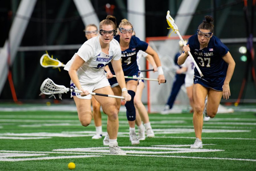 An athlete in a white jersey runs toward a lacrosse ball that sits on the ground.