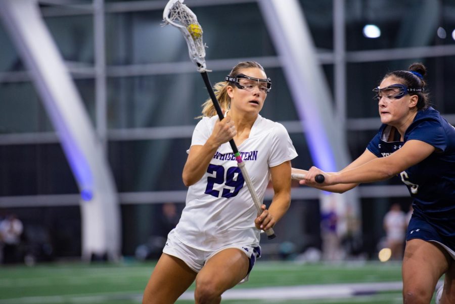 An athlete in a white jersey runs carrying a lacrosse stick, while an athlete in a blue jersey holds a lacrosse stick and tries to stop her.
