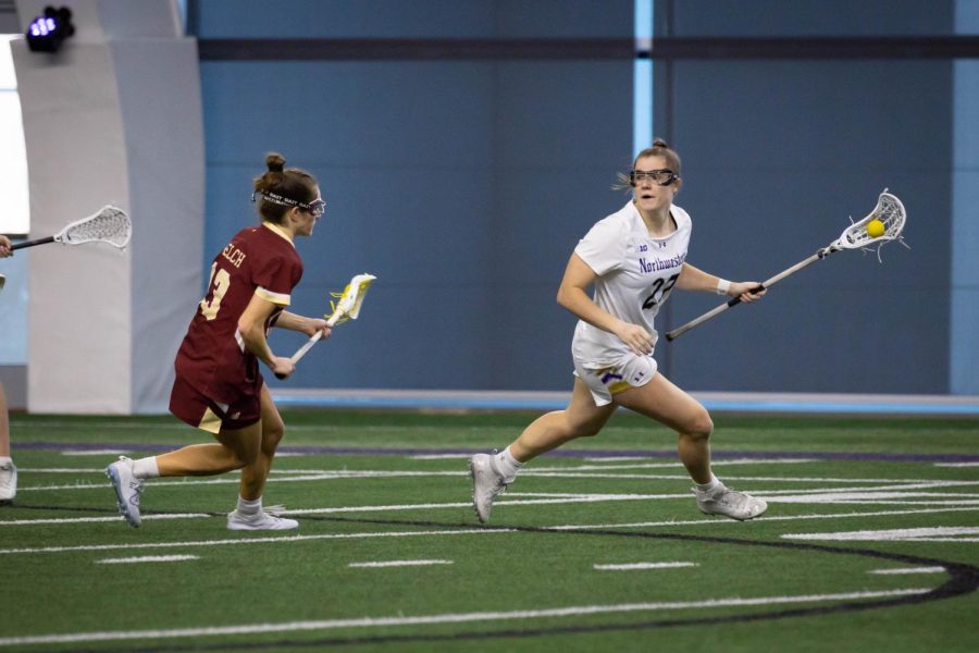 An athlete in a white jersey runs carrying a lacrosse stick.