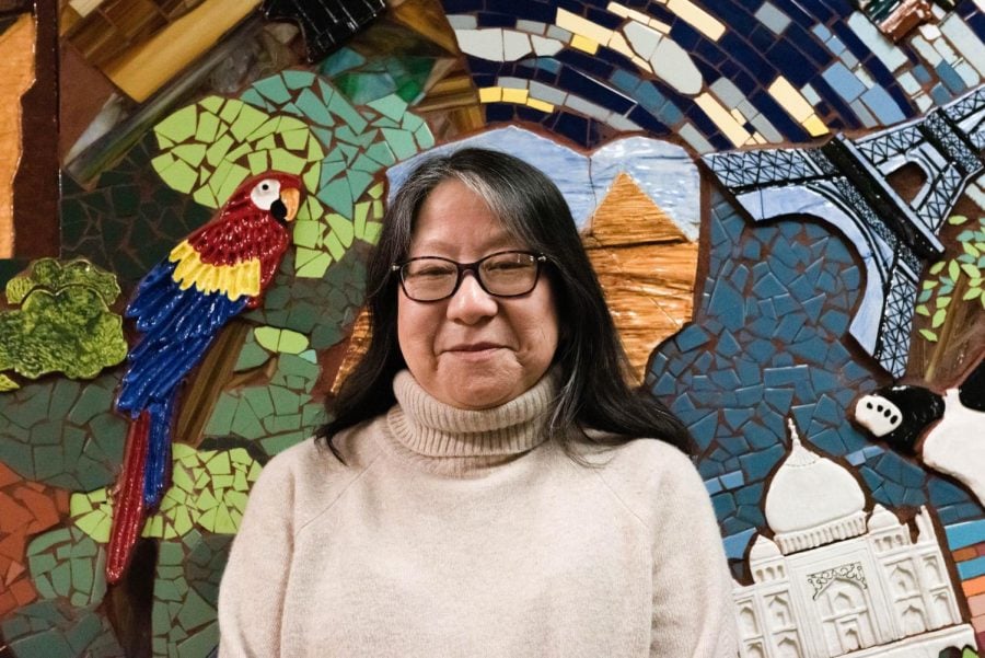 A woman wearing a tan sweater and glasses stands in front of a mosaic with a parrot.