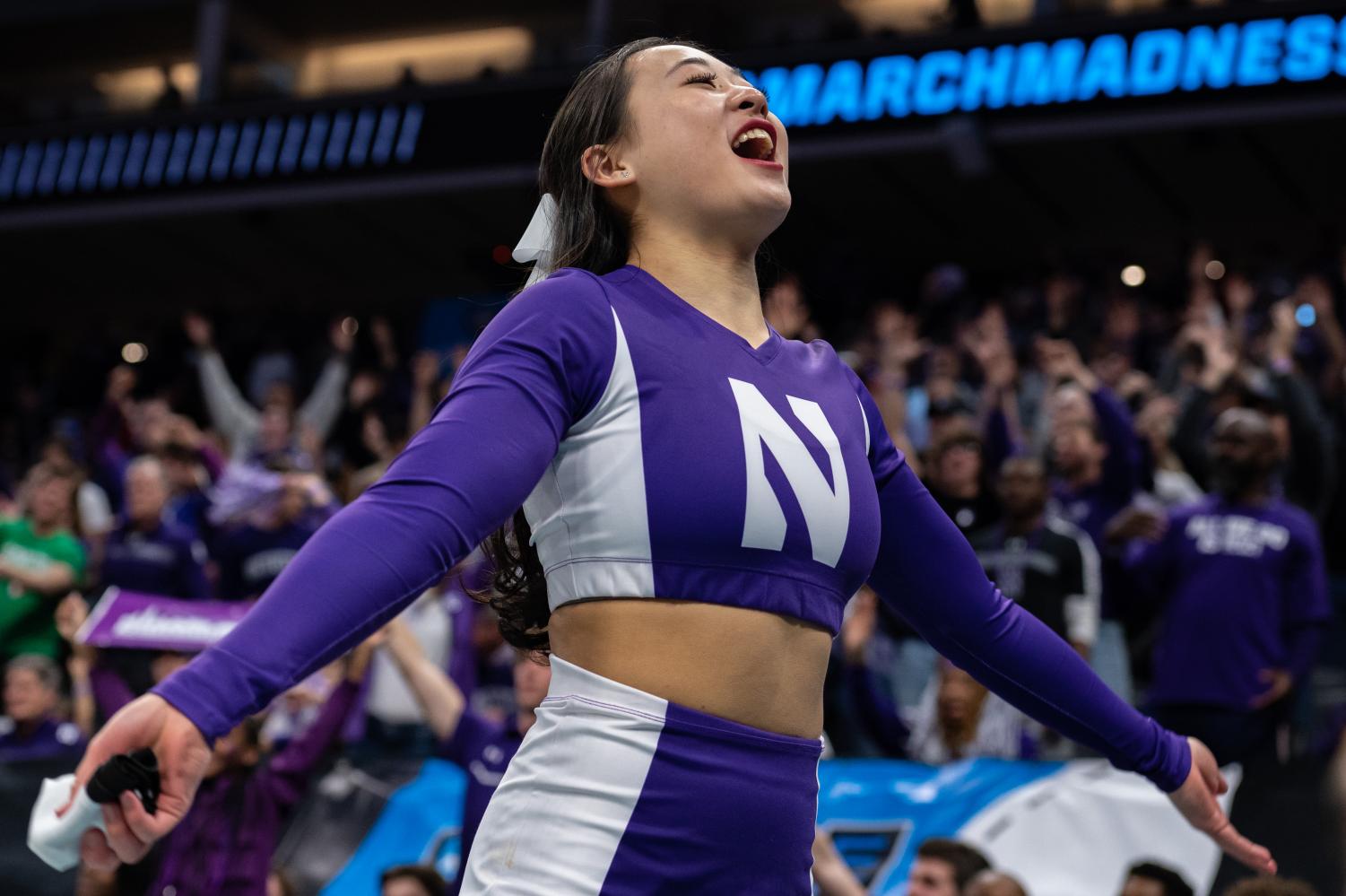 A cheerleader in purple and white spreads their arms and looks toward the crowd.
