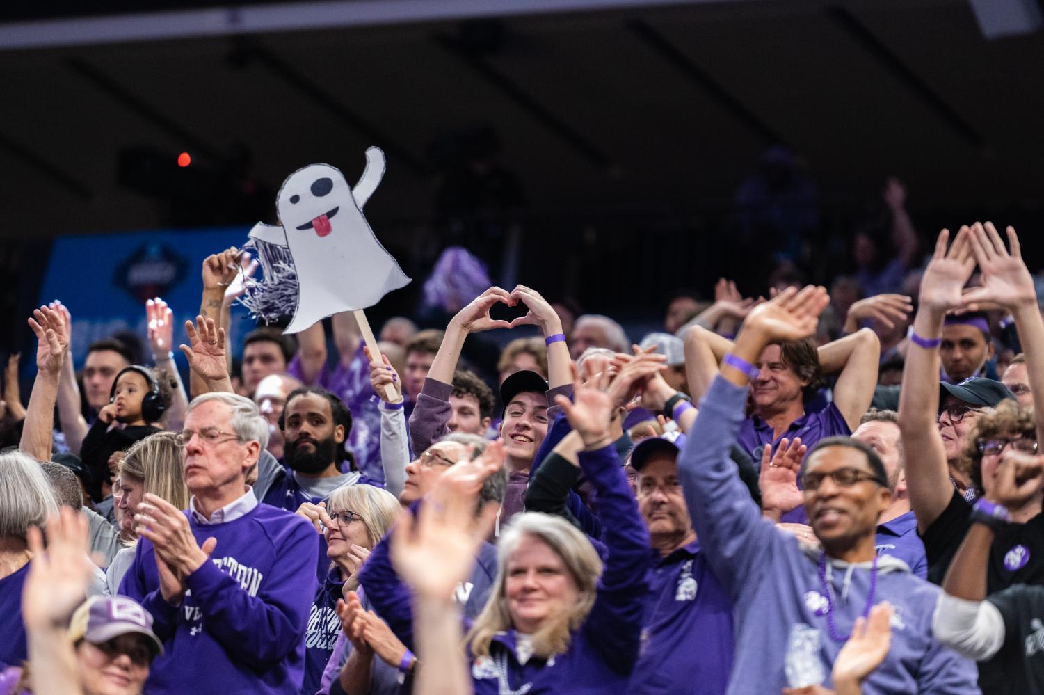 A crowd, mostly dressed in purple, cheers from the stands. One audience member makes a heart with their hands.