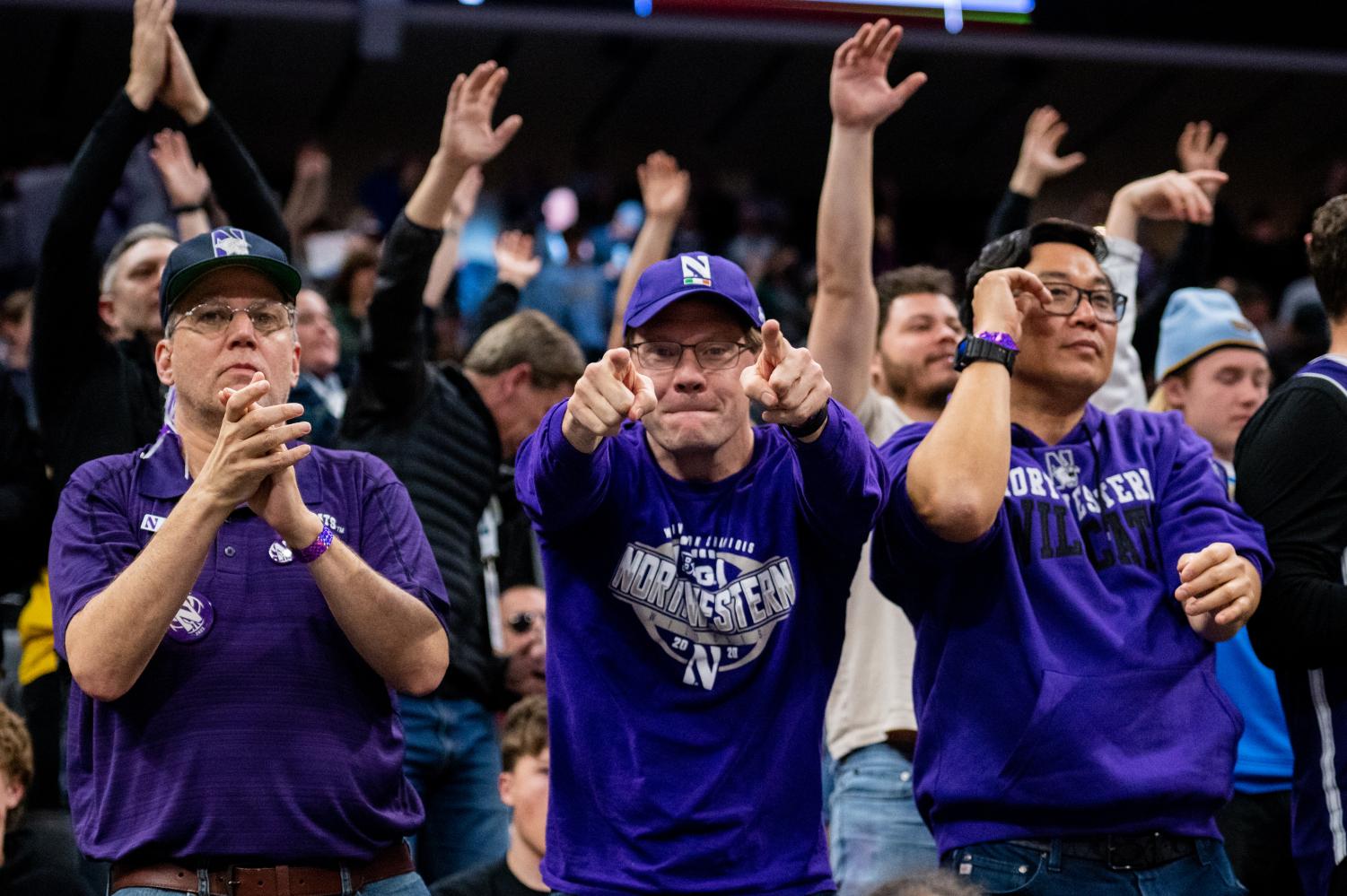 Three fans in purple cheer from the crowd. The one in the middle points to the camera with two fingers.