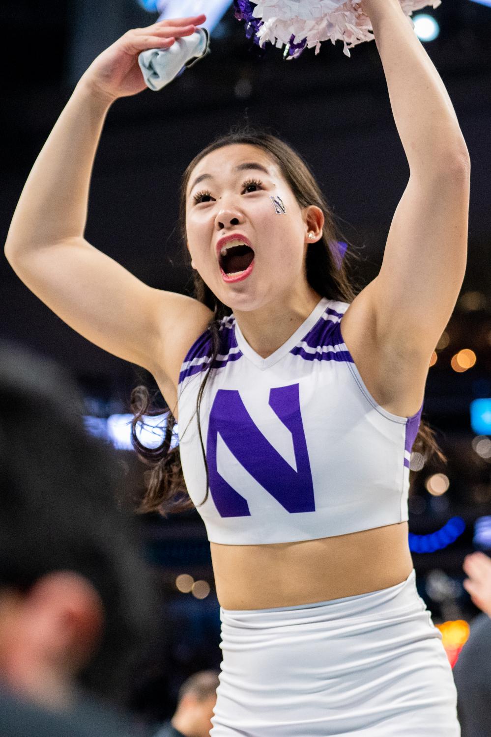 A cheerleader in white-and-purple raises their arms above their head and speaks to the crowd.
