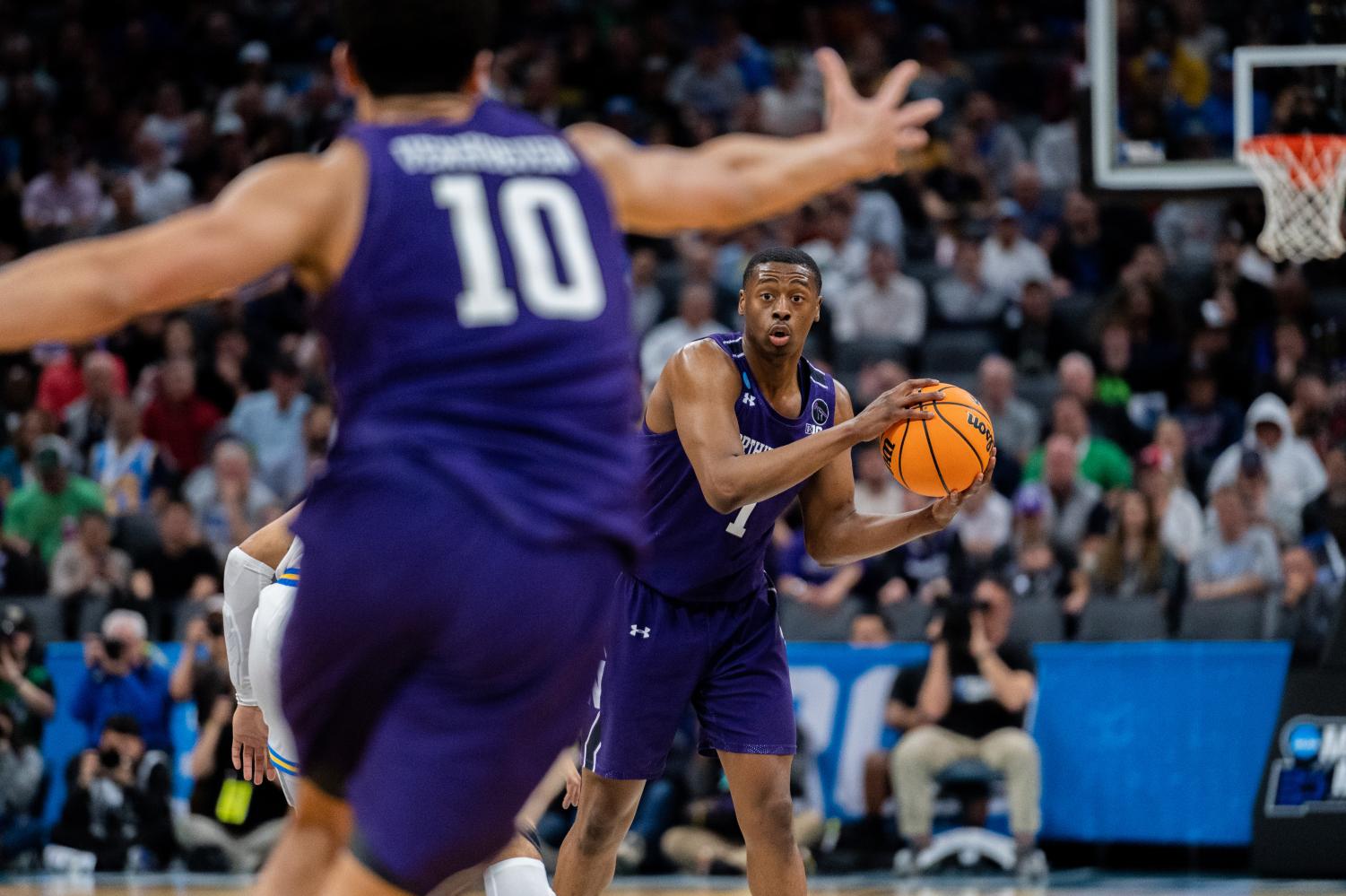A basketball player in a purple jersey passes the ball to a player in a purple jersey in front of them.