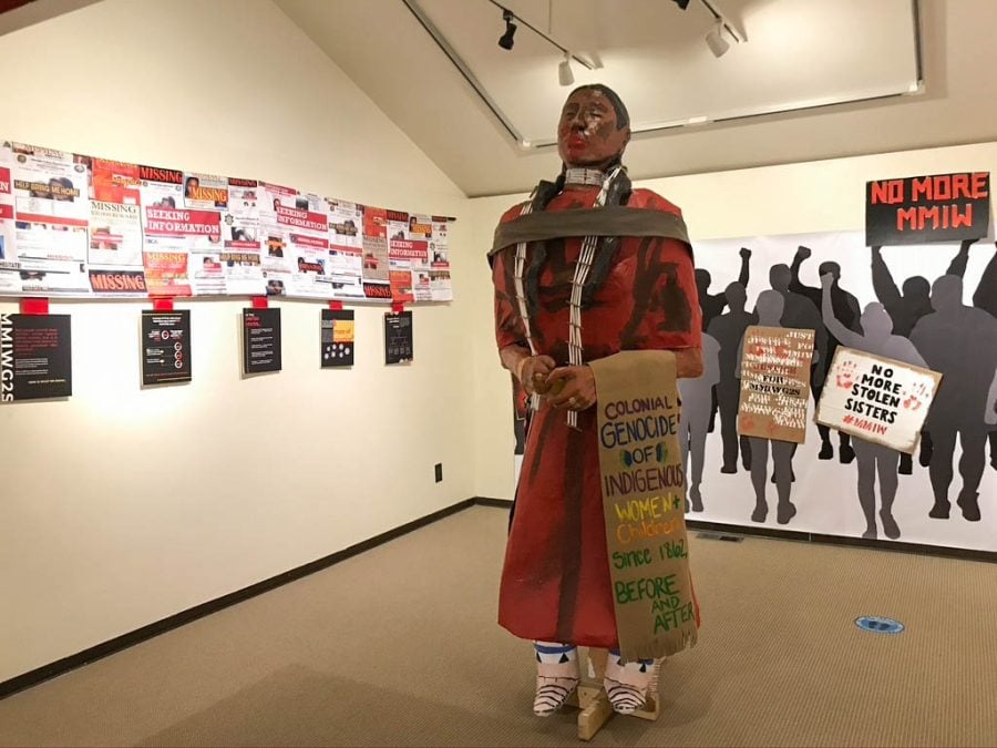 A larger-than-life statue of a person wearing red holds a banner reading, “Colonial genocide of Indigenous women + children since 1862, before and after.”