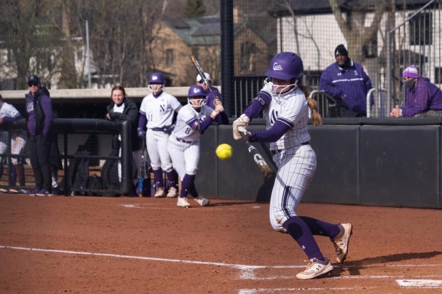 Softball player in striped purple and white uniform gears up to hit a ball with a bat.