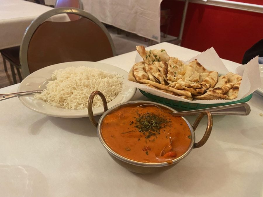 A green basket of garlic naan, a white bowl filled with white rice and a small, silver pot filled with butter chicken sit on a white tablecloth.
