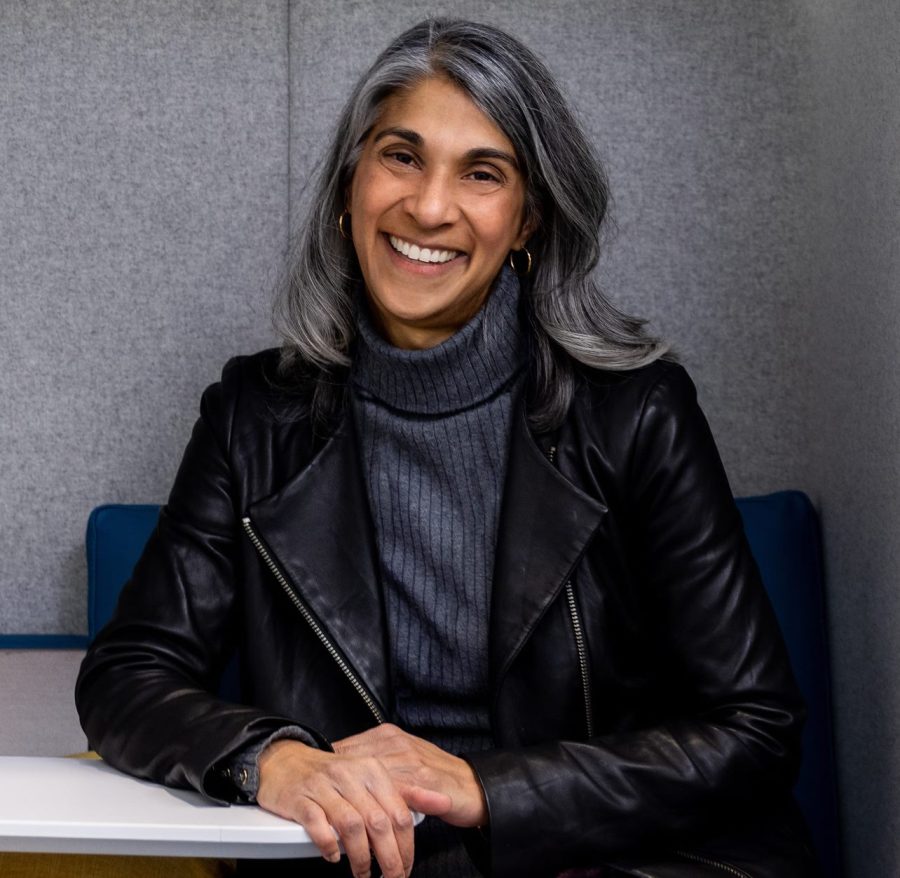 A headshot of a smiling woman with silver hair, wearing a dark-gray sweater and a black leather jacket, sitting in a blue chair. The background is a gray wall.