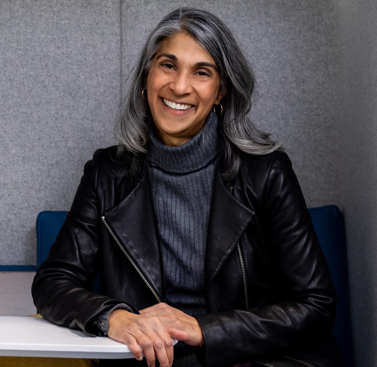 A+headshot+of+a+smiling+woman+with+silver+hair%2C+wearing+a+dark-gray+sweater+and+a+black+leather+jacket%2C+sitting+in+a+blue+chair.+The+background+is+a+gray+wall.
