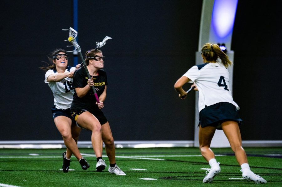 A lacrosse player in a black jersey keeps the ball away from two players in white jerseys.