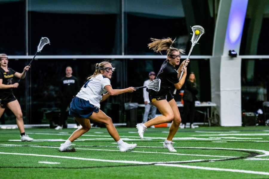 A lacrosse player in a black jersey keeps the ball away from a player in a white jersey.