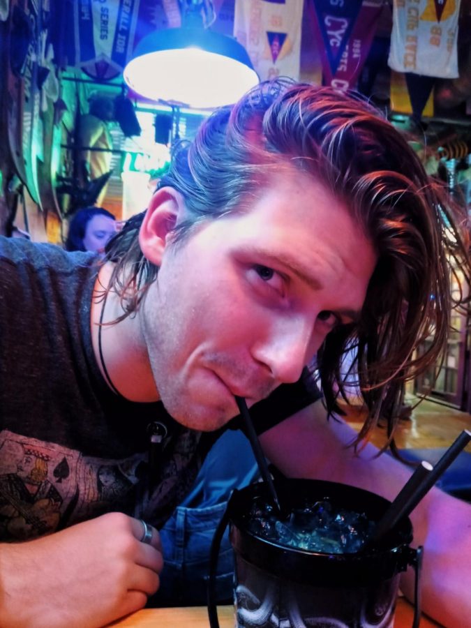 A person with medium long hair and a black t-shirt sips a drink from a straw.