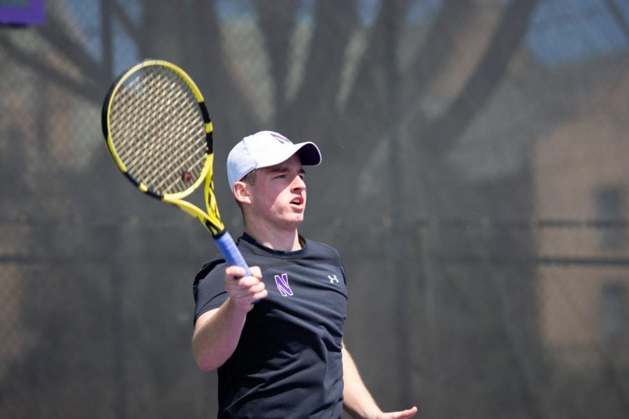 A tennis player in a black shirt and a white hat lifts up his racket.