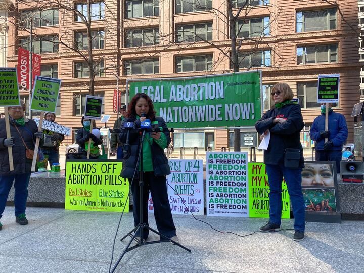 Woman speaking in front of microphones, standing in front of a sign that says “Legal abortion nationwide now.”