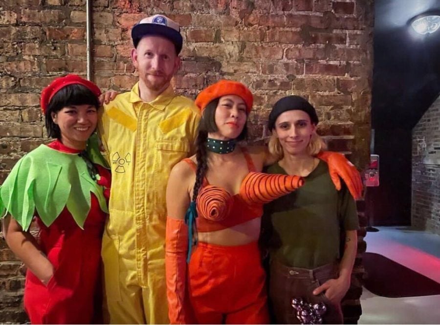 Standing in front of a brick wall are four individuals with their arms around each other. On the far left someone wears a costume in red. In the center left a person wears a yellow radiation suit and a hat. In the center right a person wears an orange beret and a large cone bra. On the right a person wears a beanie and a green shirt.