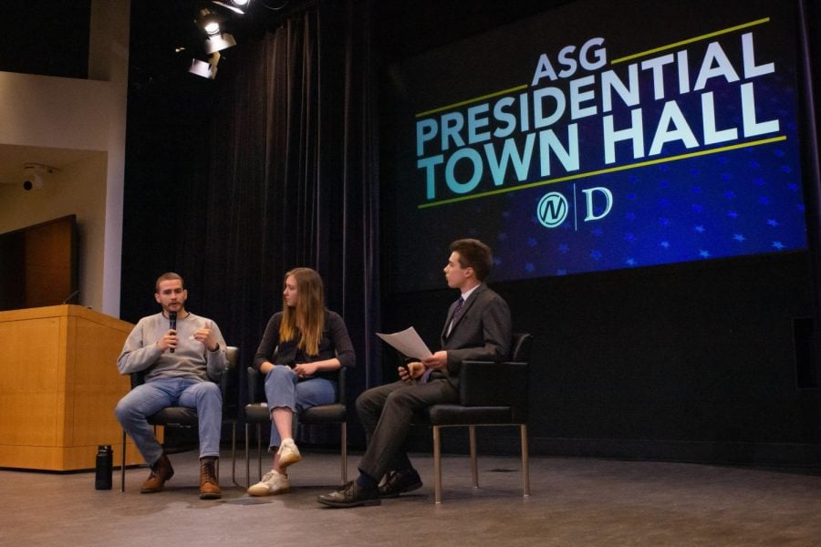 Three seated students talk to one another in a semicircle on a stage with an “ASG Presidential Town Hall” graphic in the background.
