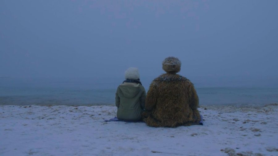 A photograph of the backs of two people sitting down next to each other in furry coats, facing a body of water in the snow.