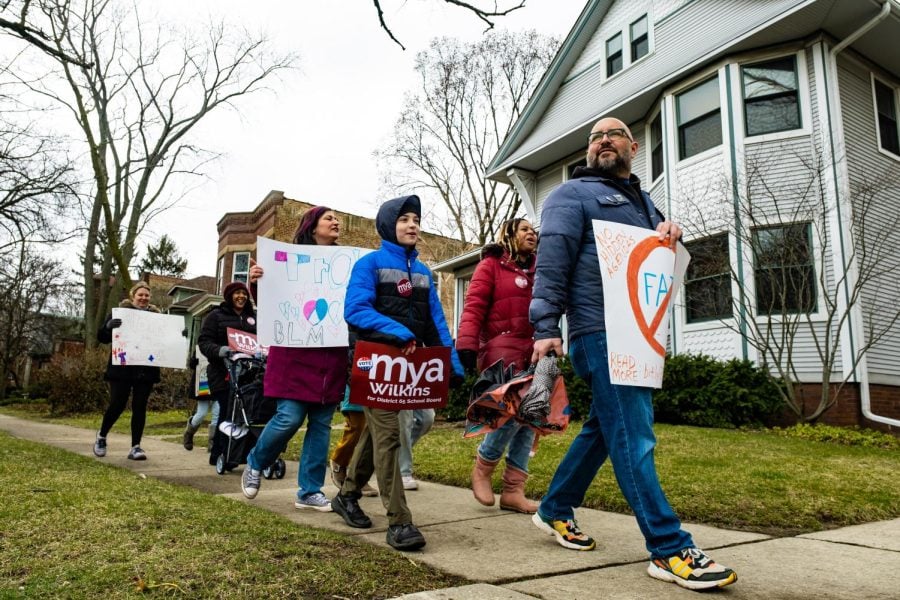 A group of people walk down the street holding signs for different D65 school board members.