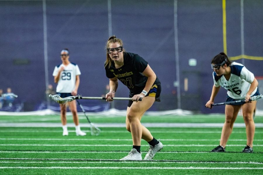 An athlete in a black jersey bends down with a lacrosse stick in her hands.