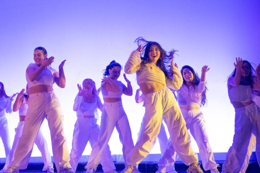 Dancers in white tops and pants perform in front of violet lights. Their forearms are up in the air, along with a dancer’s hair in motion.