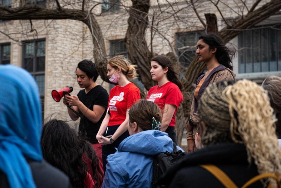 Four people stand speaking to a crowd. The person furthest left wears a black shirt and holds a red megaphone. The two people in the middle wear red shirts and the person furthest right dons a brown jacket.