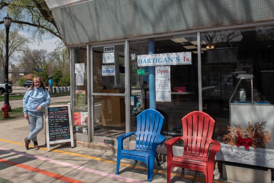 a woman leaning on a blackboard sign outside a storefront with a sign that reads “Hartigan’s Is.” The building has glass windows and and two chairs sitting outfront, one red and one blue.