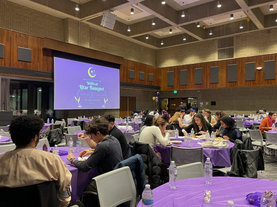 Projector screen with purple graphic that reads “Welcome To The Annual Wildcat Iftar Banquet” with purple-and-yellow embellishments. Students gathered at purple tables at the Iftar banquet.