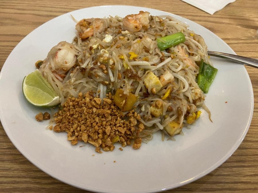 A white plate of pad thai with beige noodles, chopped peanuts, green onions, three pieces of shrimp and one slice of lime.
