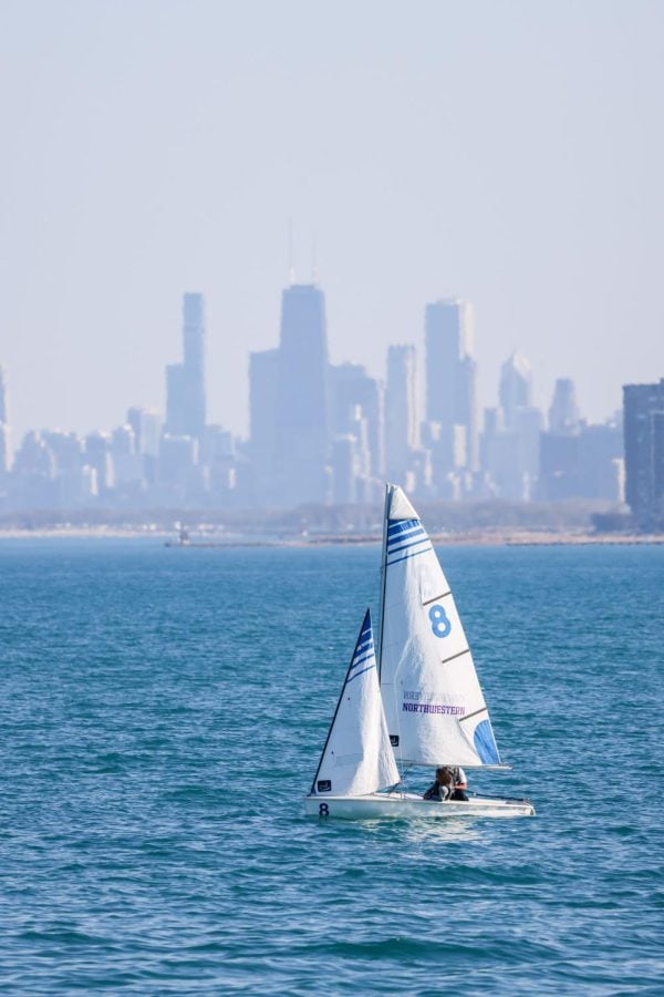 A white sailboat with a white sail with blue stripes floats on water with skyscrapers in the background.
