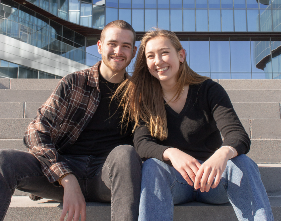 Donovan Cusick in a plaid button down, black shirt and black pants. Molly Whalen in a black long sleeve and blue jeans. The two are sitting on concrete stairs and Cusick has his hand on Whalens shoulder.