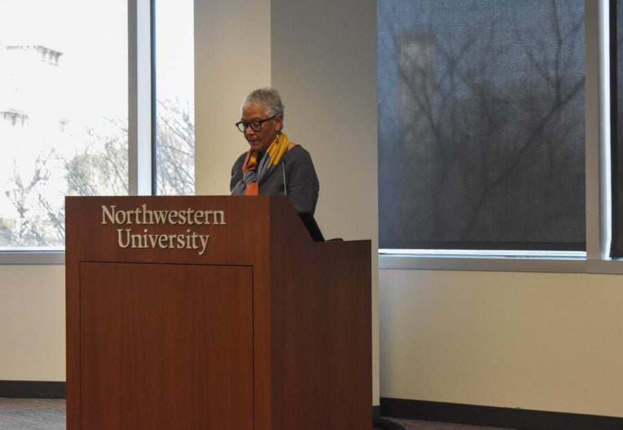 A woman in a grey sweater and glasses speaks at a brown lectern.