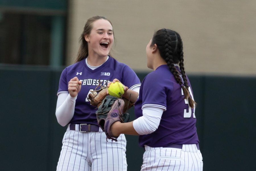 Two players in purple and white softball uniforms celebrate with smiles on their faces.