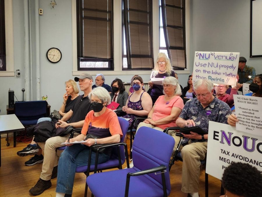 People sitting in a room, some of whom are watching a person speak, hold signs that read “Enough Tax Avoidance,” “NU Chair Peter Barris is Right: Protect us from unbridled development” and “NU venue! Use NU properly on the lake = better than Ravinia!”