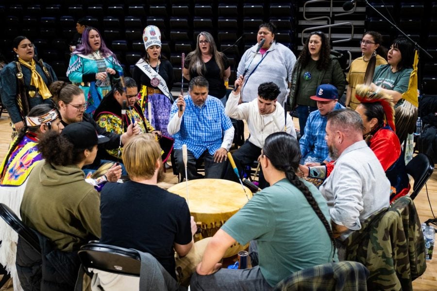 Drummers sitting in a circle and striking a drum in the middle. Behind the drummers is a group of singers.
