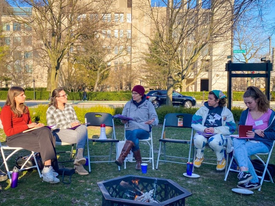 Golden light illuminates four students sitting in folding chairs and listening to a professor in a purple hat as she talks, gesturing with her hands.