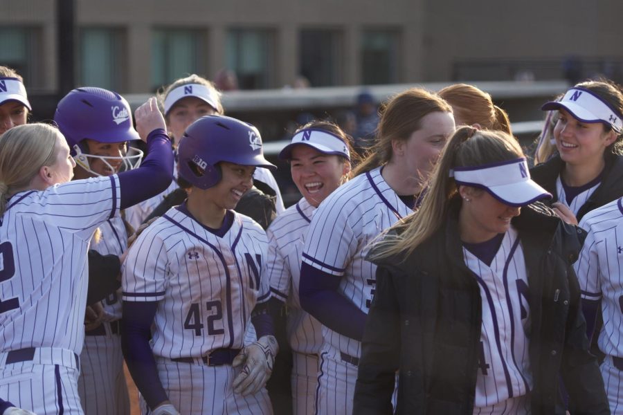 A softball player in a white jersey and white pants smiles as teammates surround them and tap their helmet.