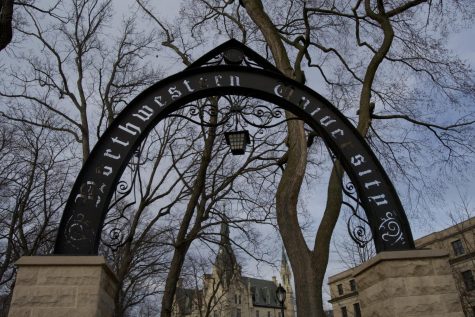 A line of barren trees behind a metal arch with “Northwestern University” inscribed on it, with a white brick building in the background.