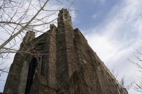 Behind barren trees, a building with a tower jutting into a blue but cloudy sky is covered in dead, brown ivy.