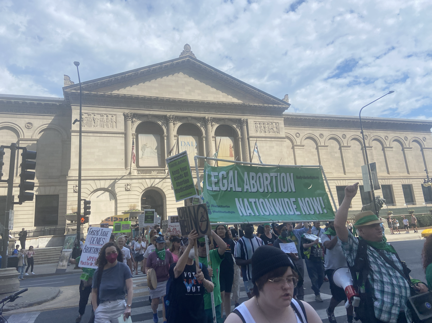 A crowd of people walk across a city street, carrying green and black signs and a green banner reading ‘Legal Abortion Nationwide.’ The Art Institute of Chicago is visible in the background. 