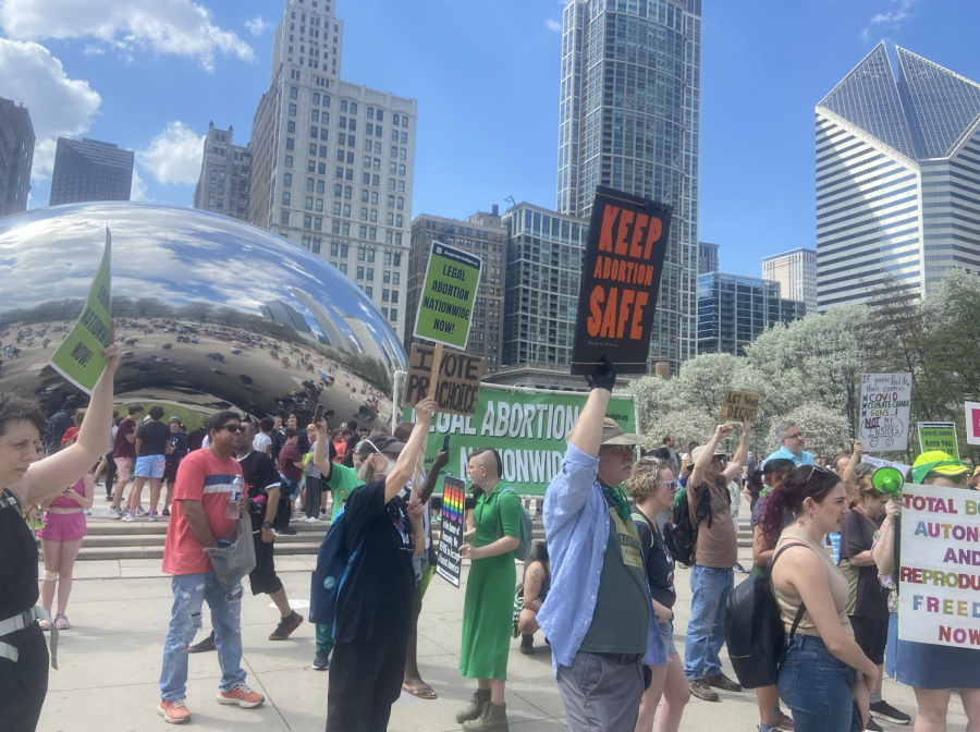 A group of people holding handmade posters stand in front of a large green banner, reading ‘Legal Abortion Nationwide.’ In the background is the Cloud Gate sculpture and the Chicago skyline.