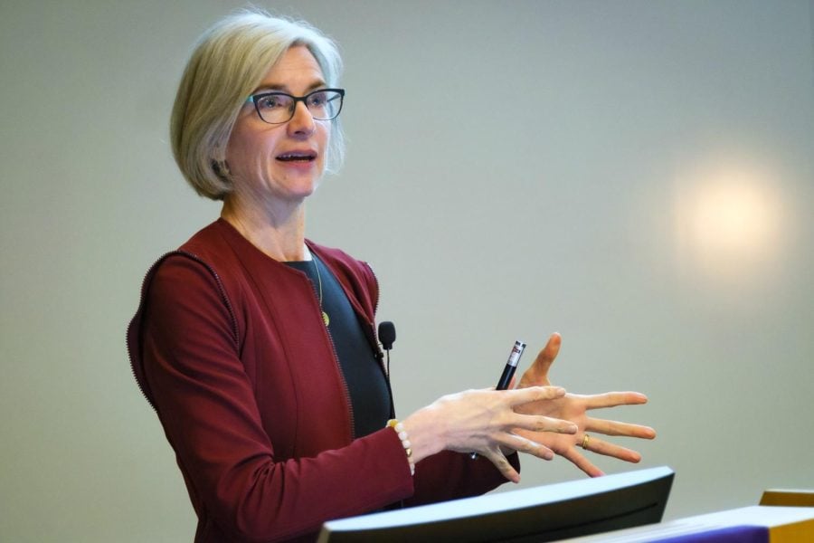 Jennifer Doudna with a short, whitish-blond bob wearing a red cardigan and black top standing at a lectern.