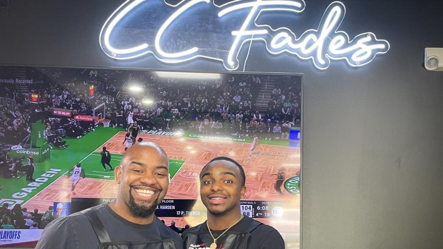 Two men stand in front of a TV under a neon sign that says “CC Fades.”