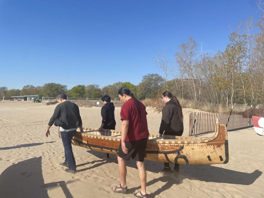 Four people are carrying a birch canoe on the beach.