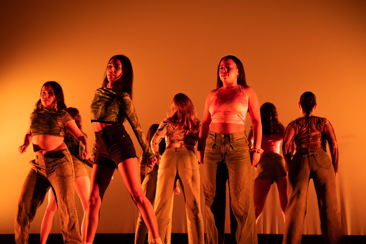 Dancers swivel their hips toward the camera in cropped shirts and denim bottoms, backlit in orange spotlights.