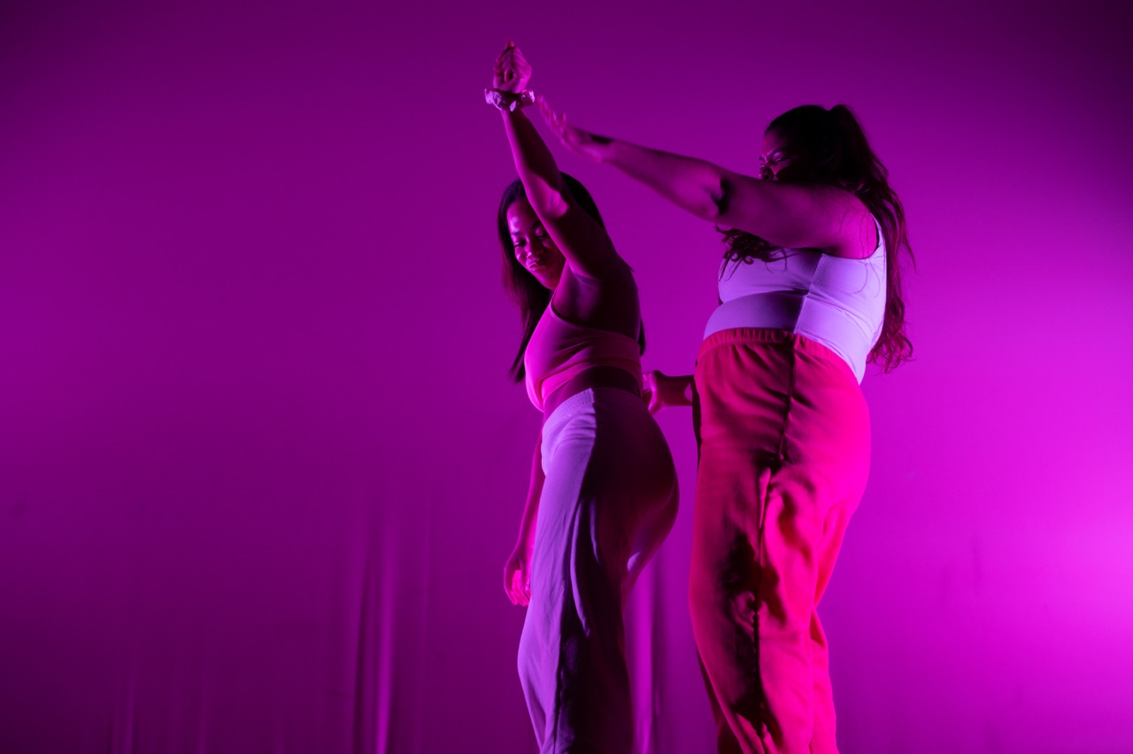 Two dancers dance together against a pink backdrop wearing sweatpants and tank tops.
